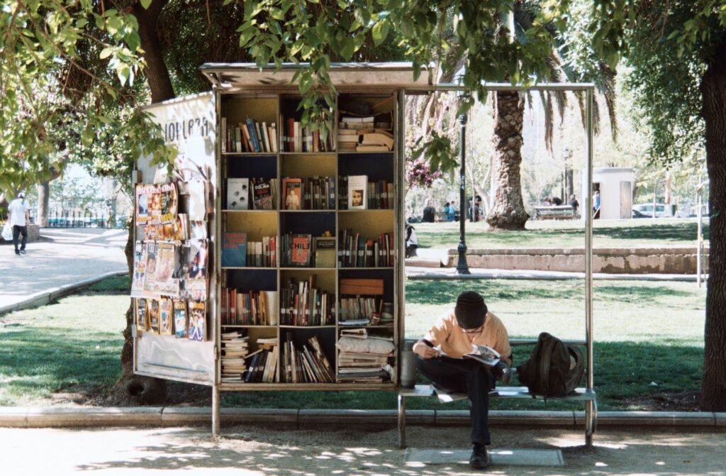 A person sitting and reading a book next to a book stall ina park